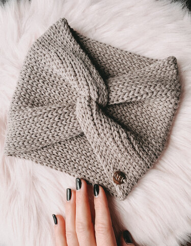 Grau gehäkeltes Stirnband mit Twistknoten auf hellem Fell, Hand mit schwarzen Nägeln berührt es