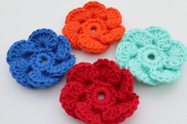 Vier gehäkelte Blumen-Applikationen in Blau, Orange, Rot und Mintgrün auf weißem Hintergrund.