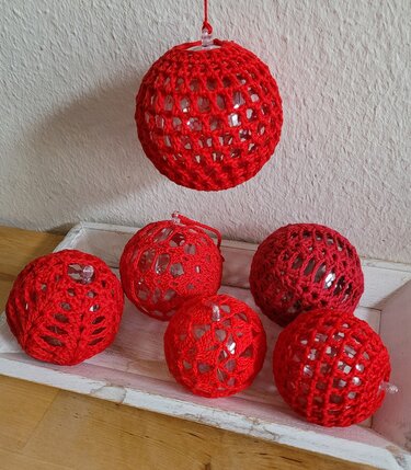 Sechs rote gehäkelte Kugeln mit transparenten Perlen in einem Netzmuster. Eine Kugel hängt, die anderen liegen in einer weißen Schale.