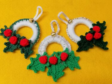 Three crochet mini holly wreath ornaments with red berries and tiny bells on yellow background