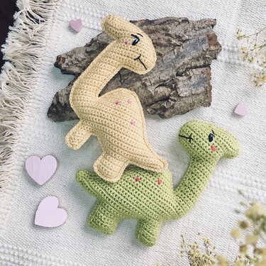 Two amigurumi dinosaur dolls, one beige and one green, on a textured white blanket with small pink hearts and a piece of wood.