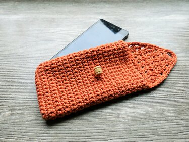 Terrakottafarbene gehäkelte Handytasche mit Holzknopf und teilweise darin steckendem Smartphone auf Holzoberfläche