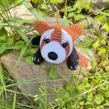 Gehäkelter roter Panda-Amigurumi mit weißem Gesicht und schwarzen Pfoten auf einem Stein im Gras