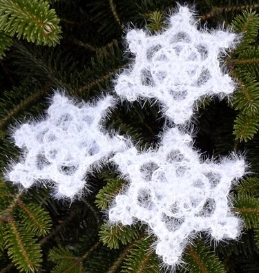 Drei weiße, flauschige Schneeflocken aus Häkelgarn auf grünen Nadelbaumzweigen