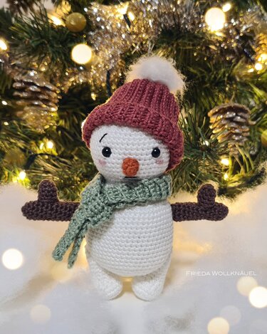 Gehäkelter Schneemann mit roter Mütze und hellgrünem Schal vor beleuchtetem Weihnachtsbaum.