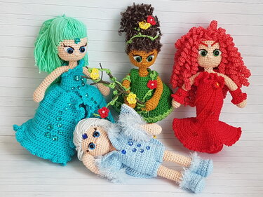 Vier gehäkelte Amigurumi-Puppen mit großen Augen und Kleidern in Türkis, Grün, Rot und Hellblau, wovon eine Puppe im Vordergrund liegt