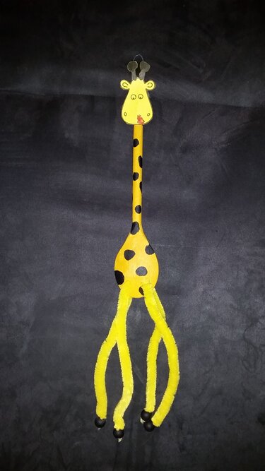 Handgebastelte Giraffe aus gelbem Holzlöffel mit schwarzen Punkten, Pfeifenreinigern als Beine und Pappkopf vor dunklem Hintergrund