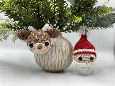 Two crocheted ornaments, Santa Claus head and reindeer head, hanging from green pine sprigs in a vase.