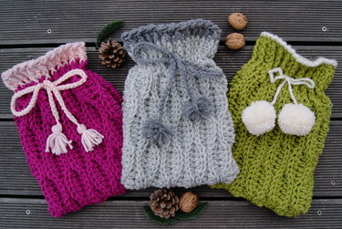Eine Reihe von gehäkelten Wärmflaschenhüllen in Pink, Grau und Grün mit unterschiedlichen Kordelzug-Designs und Zierelementen.