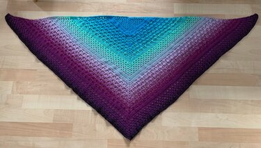 Large crocheted triangle shawl with a gradient from deep purple to teal and blue, showing textured stitch patterns, laid on a light wooden floor.