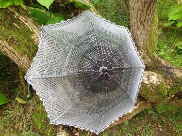 Gehäkelter Sonnenschirm in Grau-Weiß mit Spitzenmuster auf moosigem Baumstamm