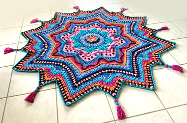 Bunte sternförmige Häkeldecke mit Mandala-Muster und pinken Quasten auf hellem Fliesenboden