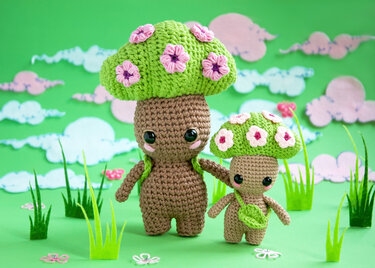 Two crocheted amigurumi mushroom dolls with green caps decorated with pink flowers on a green backdrop with felt grass.