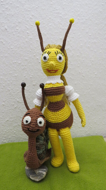 Gehäkelte gelb-braun gestreifte Amigurumi-Biene mit weißem Kragen und Puffärmeln neben brauner Amigurumi-Schnecke auf grünem Untergrund
