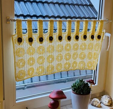 Kurz gehäkelte gelb-weiße Scheibengardine mit Reihen runder Motive und Laschen mit dunklen Knöpfen an einer Gardinenstange