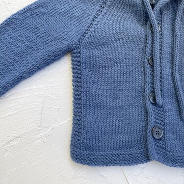 Nahaufnahme einer blauen Babyjacke mit Knopfleiste, glatt-rechts-Muster und Raglan-Ärmelansatz