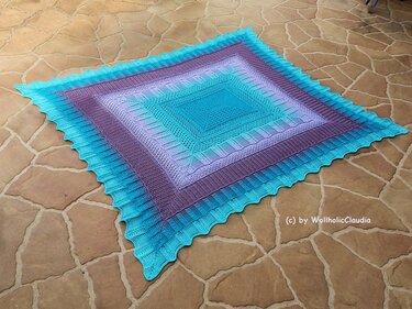 Quadratische gehäkelte Decke mit Farbverlauf von Türkis zu Lila auf Steinboden, mit gewelltem Rand