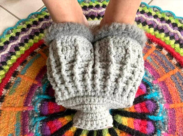 Hellgraue gehäkelte Hausschuhe mit Rippstruktur und flauschigem Rand auf buntem Häkelteppich