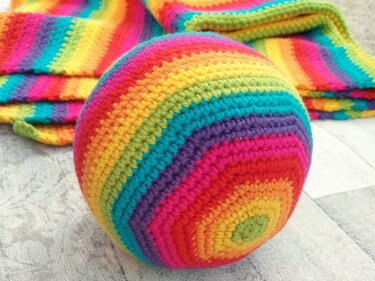 Bunter gehäkelter Ball mit Regenbogenstreifen vor gestreifter Häkeldecke