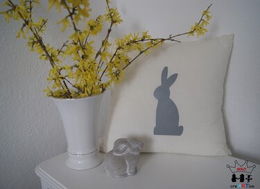 Kissen mit geplottetem Hasenmotiv, daneben gelbe Forsythien in einer Vase und eine kleine Hasenfigur.