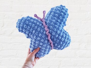 Hand holding a blue crocheted butterfly-shaped pillow with popcorn bobble texture and a purple twisted body