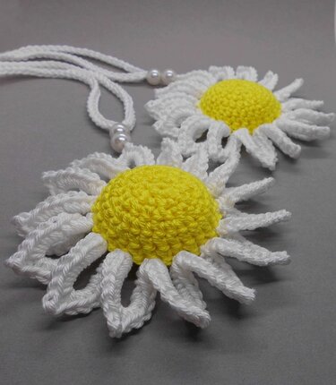 Two crocheted daisy motifs with yellow centers and white looped petals on a cord with round beads, on a gray background