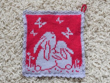 Red knitted square with two light lavender rabbits embracing, four butterflies above, scalloped lavender border, red hanging loop, on a cream shaggy rug.