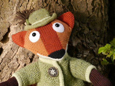 Hand-knitted fox amigurumi wearing a moss-green jacket and small felt hat, leaning against tree bark