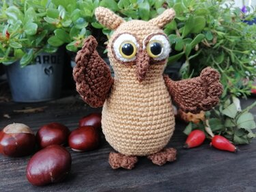 Braune gehäkelte Amigurumi-Eule mit großen Knopfaugen vor Pflanzen und Kastanien
