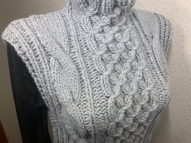 Close-up of a light gray sleeveless turtleneck vest with cable and basketweave stitch patterns.