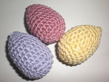 Drei handgehäkelte Ostereier in Pastellfarben (lila, rosa, gelb) auf hellem Untergrund