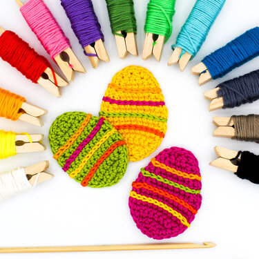 Nahaufnahme: drei gehäkelte Ostereier in leuchtenden Farben, umgeben von auf Holzklammern gewickeltem Garn und einer Häkelnadel.