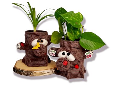 Two brown crocheted tree-stump plant pot covers with large white eyes, round snouts and potted green houseplants