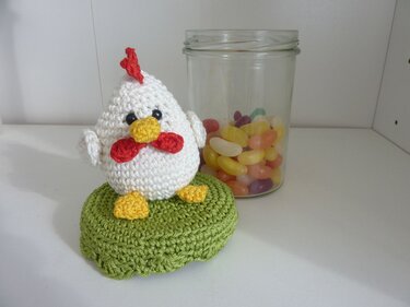 Weißes gehäkeltes Huhn mit rotem Kamm auf grünem Untergrund, neben einem Glas mit bunten Geleebohnen.