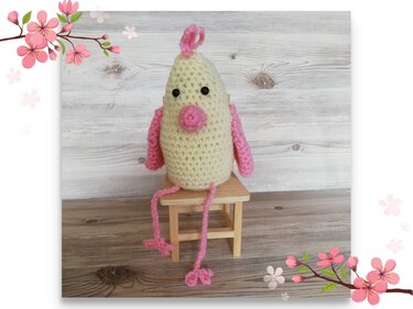 Kleines gelbes gehäkeltes Küken mit rosa Flügeln, Schnabel, Kamm und Beinen auf einem Mini-Holzhocker.