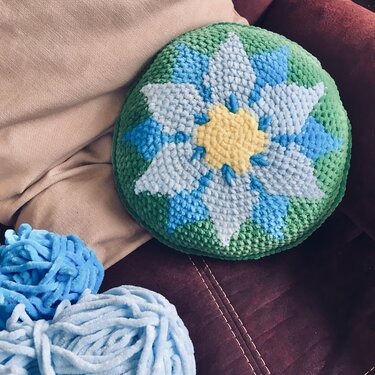 Rundes gehäkeltes Kissen mit blau-weißem Blumenstern auf grünem Hintergrund, hellblaue Wollknäuel daneben, auf Sofa