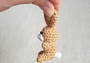 Beiger gehäkelter Mini-Hase mit weißem Schwänzchen, Seitenansicht, an Ohrspitze gehalten