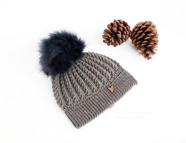 Gehäkelte graue Mütze mit Rippstich, breitem Bündchen, dunklem Bommel und herzförmigem Knopf neben Kiefernzapfen