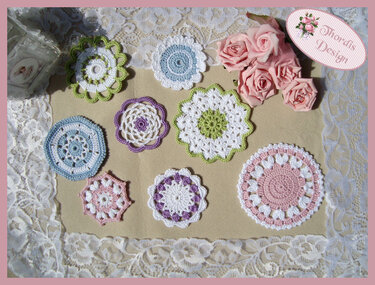 Mehrere runde gehäkelte Applikationen in Pastellfarben auf Spitzenstoff, daneben rosafarbene Rosen