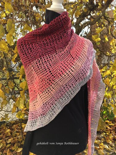 Gehäkeltes Dreieckstuch in Rot-, Rosa- und Grautönen mit Farbverlauf und Lochmuster, drapiert an einer Schaufensterpuppe vor herbstlichem Laub.
