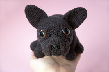 Hand holding a small dark crocheted amigurumi dog with large plastic safety eyes and upright ears on a pink background