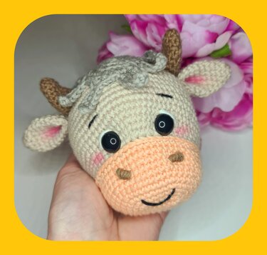 Hand hält gehäkelten Amigurumi-Kuhkopf in Pastellfarben mit schwarzen Augen und rosa Wangen
