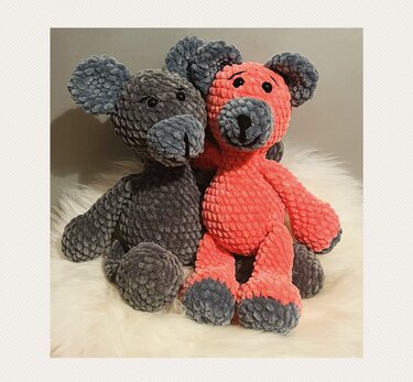 Zwei gehäkelte Amigurumi-Teddys aus samtigem Plüschgarn, einer grau, einer korallenrot mit grauen Ohren und Pfoten, sitzend auf weißem Fell