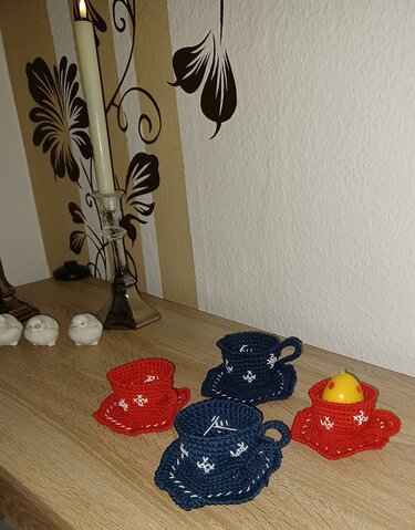 Gehäkelte Tassenuntersetzer in Rot und Dunkelblau mit weißen Schneeflocken auf einem Holztisch, Kerze und Dekoobjekte im Hintergrund.