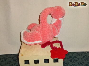 Rosa, handgehäkeltes Flamingo-Kuscheltier mit weißen Rändern an den Flügeln, auf einer hellen Holzbox, daneben ein rotes Beißspielzeug.