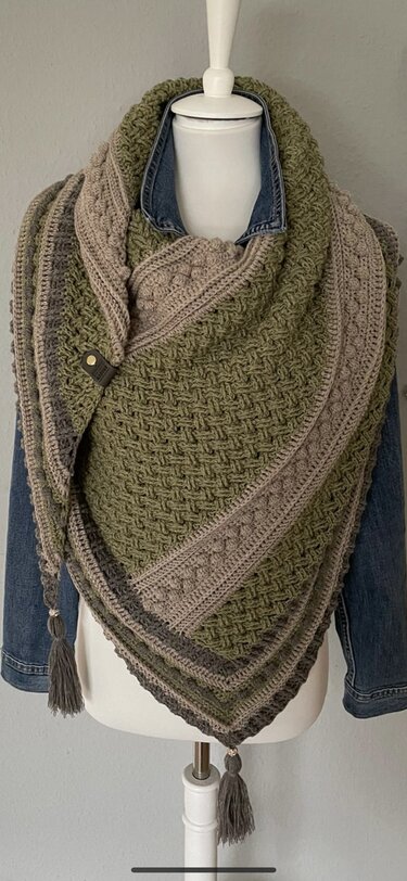 Gehäkeltes Dreieckstuch in Oliv, Beige und Grau mit strukturiertem Muster, Quasten und Verschluss auf einer Schneiderpuppe.