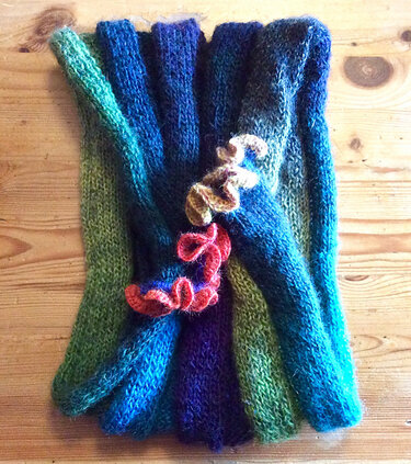 Knitted loop scarf in green-to-blue gradient with reddish-orange and light brown ruffles on a wooden surface.