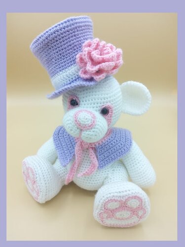 Weißer Amigurumi-Teddybär mit lila Zylinderhut und rosa Rose, sitzend