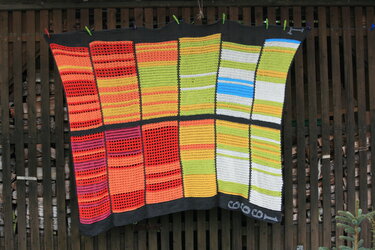 Bunte gehäkelte Patchwork-Decke mit verschiedenen Farbblöcken und Lochmustern, aufgehängt an einer Leine.