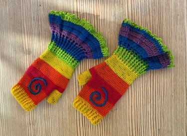 Gehäkelte fingerlose Armstulpen in Regenbogenfarben mit blauem Spiralmuster auf hellem Holzuntergrund.